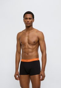 Μαύρα ανδρικά εσώρουχα τύπου μποξέρ με πορτοκαλί ζώνη μέσης, με το όνομα της μάρκας "TOMMY HILFIGER". Μαλακό ύφασμα με εφαρμοστή σχεδίαση.