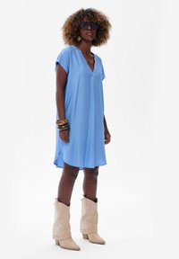 Lekka, niebieska sukienka w serek z krótkimi rękawami, charakteryzująca się luźnym krojem i bocznymi rozcięciami, zestawiona z beżowymi kowbojkami.