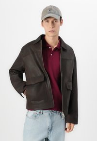 Un jeune homme portant une casquette grise, une veste en cuir marron foncé, un polo bordeaux et un jean bleu clair, se tenant devant un fond blanc.