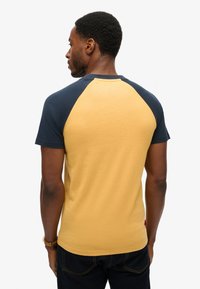 Camisa de manga corta con un cuerpo amarillo y mangas raglán en azul marino, confeccionada en suave tela de algodón y con detalles de costura en contraste.