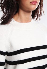 Sweter wełniany z białym korpusem i czarnymi poziomymi paskami. Posiada okrągły dekolt oraz prążkowane wykończenia przy kołnierzu i dolnym brzegu.