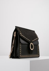 Borsa nera con un design strutturato, parte superiore in suede, dettagli con borchie dorate e una fibbia circolare sulla parte anteriore.