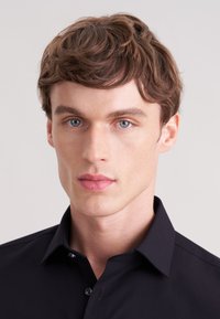 Zwart overhemd met knopen en kraag, gemaakt van effen stof met een subtiele glans. Close-up van het gezicht van een mannelijke model, met een lichte huid en kort haar.