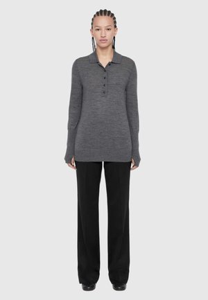 Person with braided hair wears a long-sleeve gray buttoned sweater and loose black trousers, standing against a plain light background.