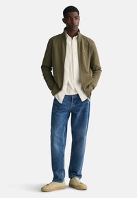 Chaqueta verde oliva con cremallera sobre una camisa blanca de botones, combinada con vaqueros azules y zapatillas beige. La chaqueta tiene bolsillos delanteros y cuello.