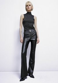 Pantaloni neri in pelle a zampa d'elefante con vita alta, dettagli di bottoni decorativi e una texture liscia. Abbinati a un top nero attillato senza maniche.