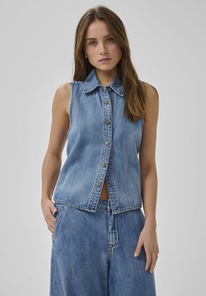 Młoda kobieta ubrana w bezrękawnik z dżinsowego materiału zapinany na guziki oraz luźne dżinsowe spodnie, stojąca z jedną ręką w kieszeni na jednolitym tle.