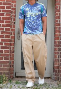 Niebieska graficzna koszulka Adidas z żółtymi akcentami i logo "Ziggo", w zestawie z beżowymi spodniami cargo i białymi trampkami. Tło stanowi ceglana ściana i drzwi.