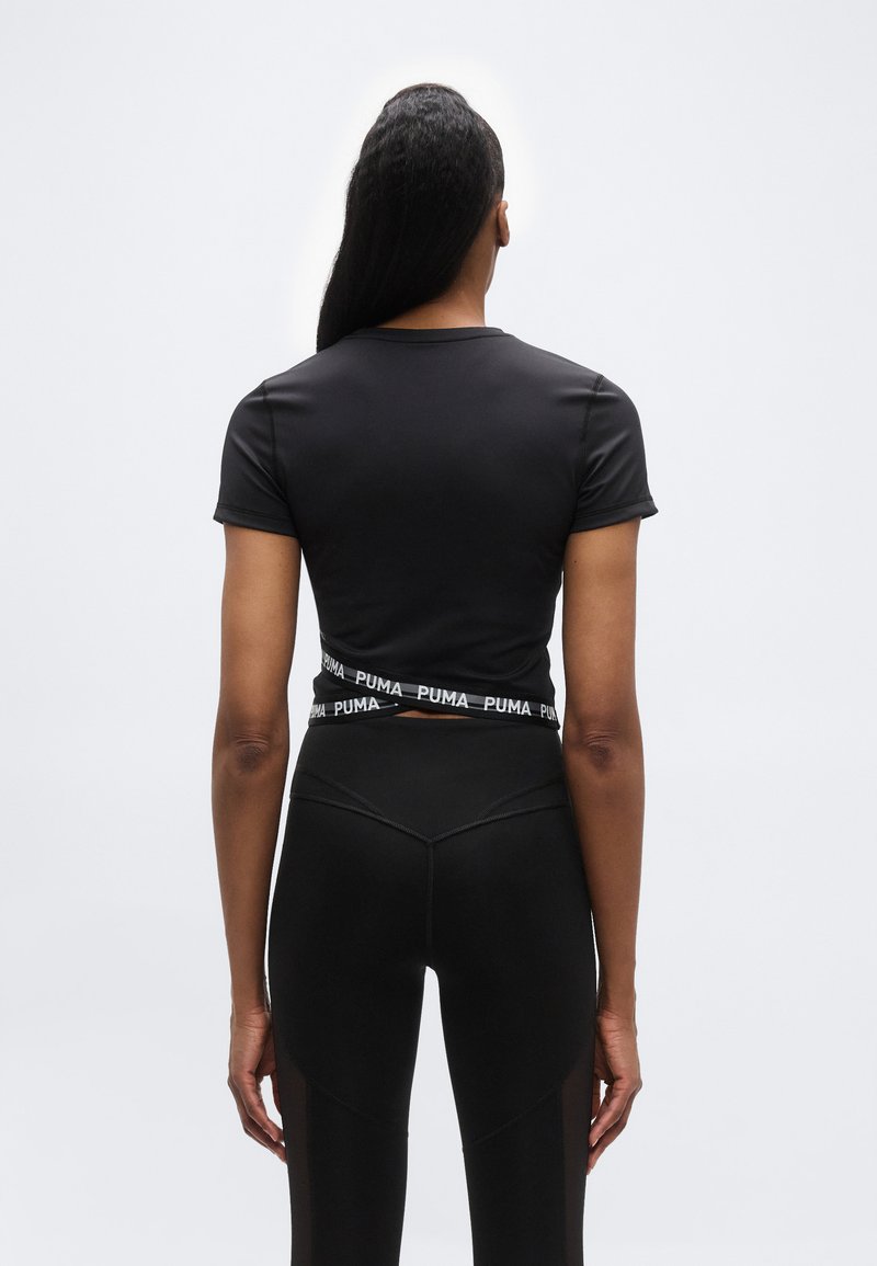 Γυναίκα με μακριά μαλλιά που φοράει μαύρη κοντομάνικη μπλούζα με το λογότυπο Puma και λεπτομέρεια τύπου wrap, καθώς και μαύρο κολάν, στέκεται γυρισμένη προς τα πίσω έχοντας λευκό φόντο.