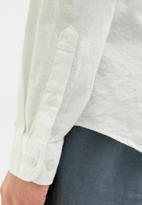 Manica di camicia in lino bianco con polsino appuntito, dotata di due bottoni chiari. Il tessuto ha una finitura leggermente ruvida. Sotto è presente un tessuto scuro.