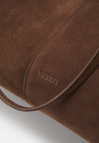 Brązowa torba z zamszu o gładkiej texture, z logo wytłoczonym na powierzchni i eleganckim, minimalistycznym designie.