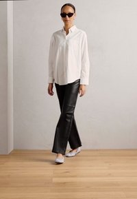 Camicia bianca a maniche lunghe, pantaloni in pelle neri a campana e calzature grigio chiaro; la modella indossa occhiali da sole neri; sfondo minimalista.