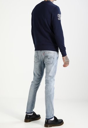 Felpa navy con colletto a girocollo e numero bianco sulla manica, abbinata a jeans azzurri chiari e scarpe nere con un leggero tacco.