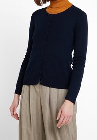 Marinblå cardigan med lång ärm och rund halsringning, med knäppning fram och ribbade manschetter, kombinerad med beige byxor med smala ränder.