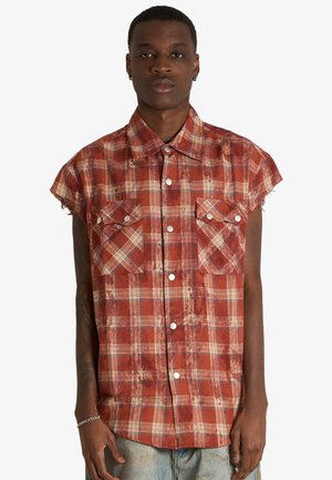Giovane uomo che indossa una camicia a quadri rossa e beige senza maniche con due tasche sul petto, in piedi davanti a uno sfondo semplice.