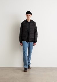 Veste rembourrée noire avec fermeture éclair, associée à une chemise noire et un jean bleu clair. Le modèle porte un bonnet noir et des chaussures marron.