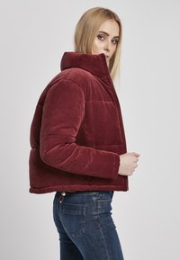 Rød kordfløjl pufferjakke med høj krave, vandret quiltet design og sidelommer, båret over mørkeblå jeans.