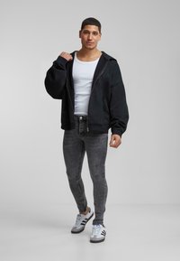 Crna zip-up dukserica s oversized rukavima, bijela majica na bretele, sive uske traperice i crno-bijele tenisice. Glatka tkanina i ležeran dizajn.