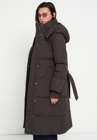 Γυναίκα που φοράει μακρύ σκούρο μπουφάν με κουκούλα και ζώνη, στέκεται πλαγίως, επίσης φοράει χρωματιστά ορθογώνια γυαλιά ηλίου, μπροστά σε απλό φόντο.