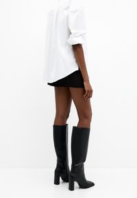 Bottes noires montantes jusqu'aux genoux avec un talon carré, une texture en cuir lisse, un bout carré, et une fermeture éclair à l'arrière, portées avec une chemise blanche.