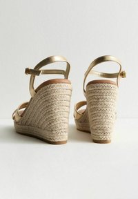 Les sandales à wedge métalliques dorées présentent une plateforme en jute tressé, une semelle intérieure lisse marron et des brides ajustables à la cheville.