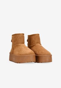 Botas de tobillo de gamuza color marrón claro con punta redondeada, suela plana de plataforma y acentos de costura. La parte trasera cuenta con un lazo para facilitar el calzado.