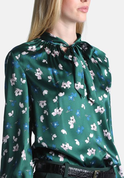 Femme portant une blouse en satin verte avec imprimé floral bleu et blanc et un nœud noué au niveau de l'encolure, associée à une ceinture noire texturée.
