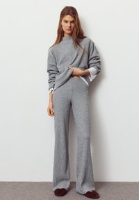 Grauer Strickpullover mit hohem Ausschnitt und Ärmel-Details, kombiniert mit passenden, weit geschnittenen Hosen. Das Modell trägt gemütliche braune Hausschuhe.