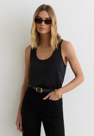 Žena s blond vlasmi po plecia nosi čierne slnečné okuliare, čierne tielko bez rukávov, čierne nohavice s opaskom a masívny zlatý náramok.