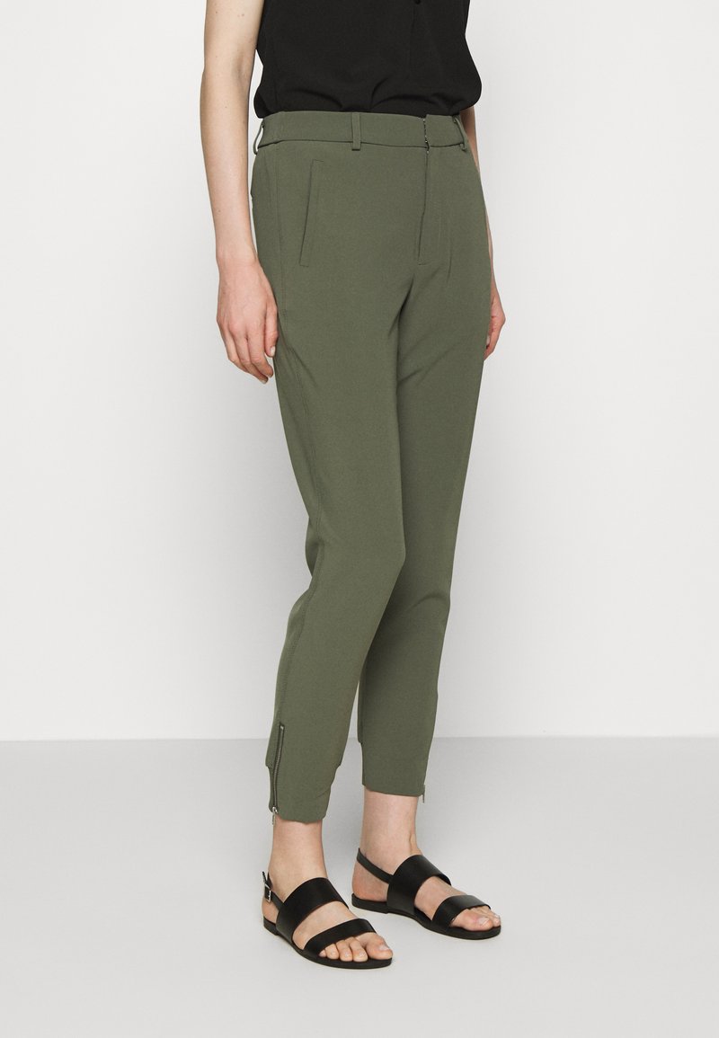 Calças ajustadas verde-azeitona com bolsos frontais e fechos de correr na bainha. Feitas de um tecido suave, combinadas com uma blusa preta e sandálias.