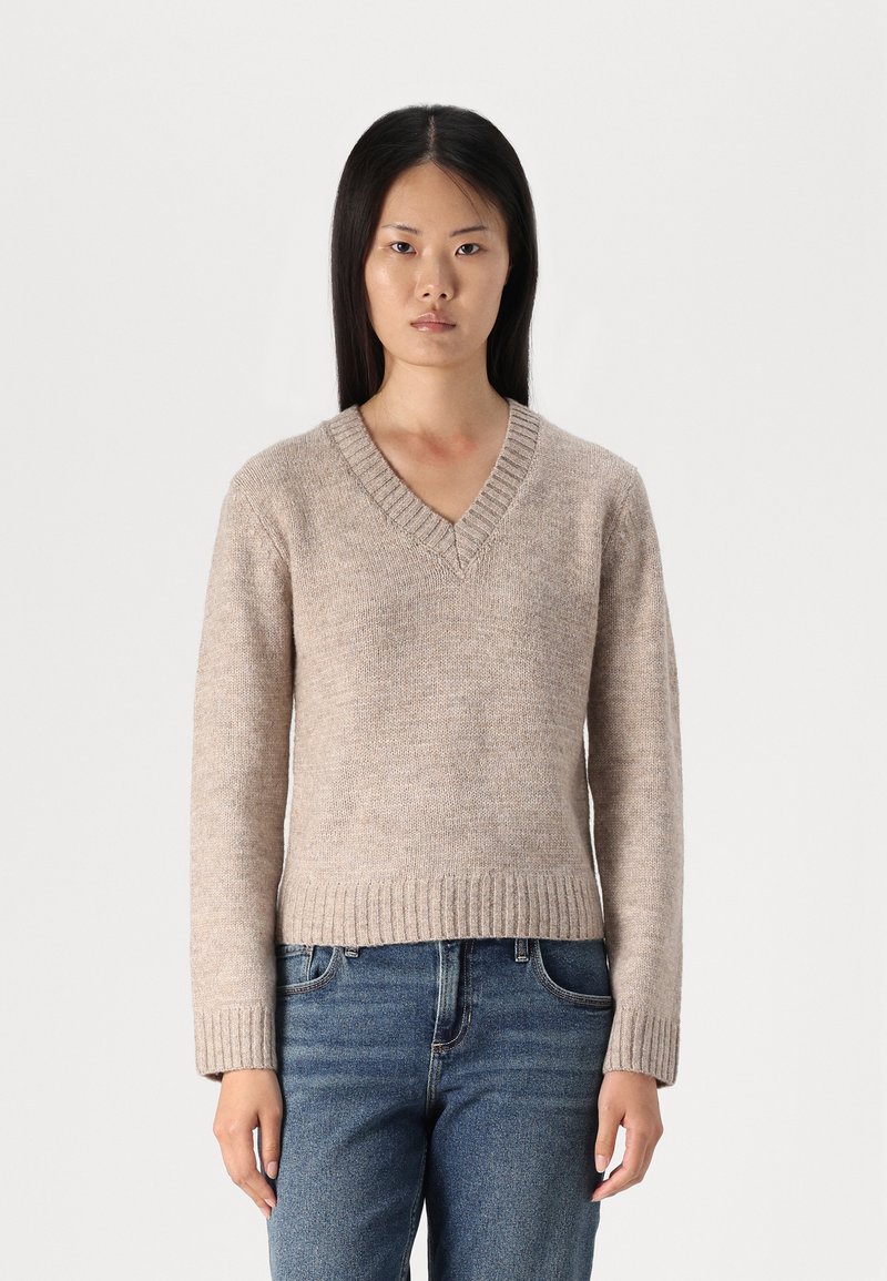 Pull à col en V beige clair en tissu tricoté texturé, doté de manches longues et d'un ourlet côtelé, porté avec un jean en denim bleu.