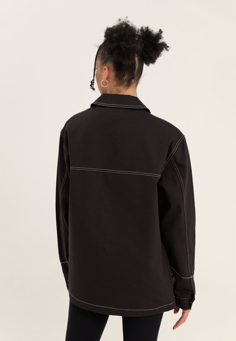 Person med afro-texturerat hår i en knut iförd en lös svart jacka med vita sömmar, stående vänd bort från kameran mot en enkel bakgrund.