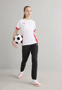 Valge spordisärk punaste detailidega, mustad püksid, hoiab musta-valget jalgpalli, kannab valgeid sportlikke jalanõusid oranžide detailidega.