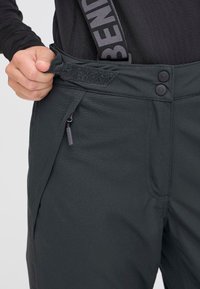 Mørk grå utendørsbukser med en glatt tekstur, som har to knapper, en borrelåsrem og en glidelåslomme på siden.