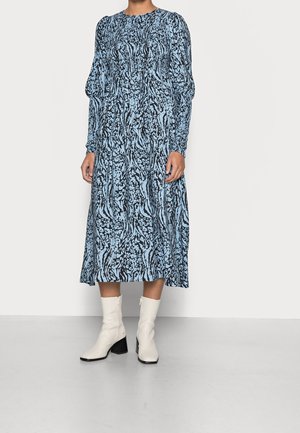 Robe longue à manches longues bleu et noire avec une taille froncée et une jupe fluide, portée avec des bottines blanches et un talon empilé.