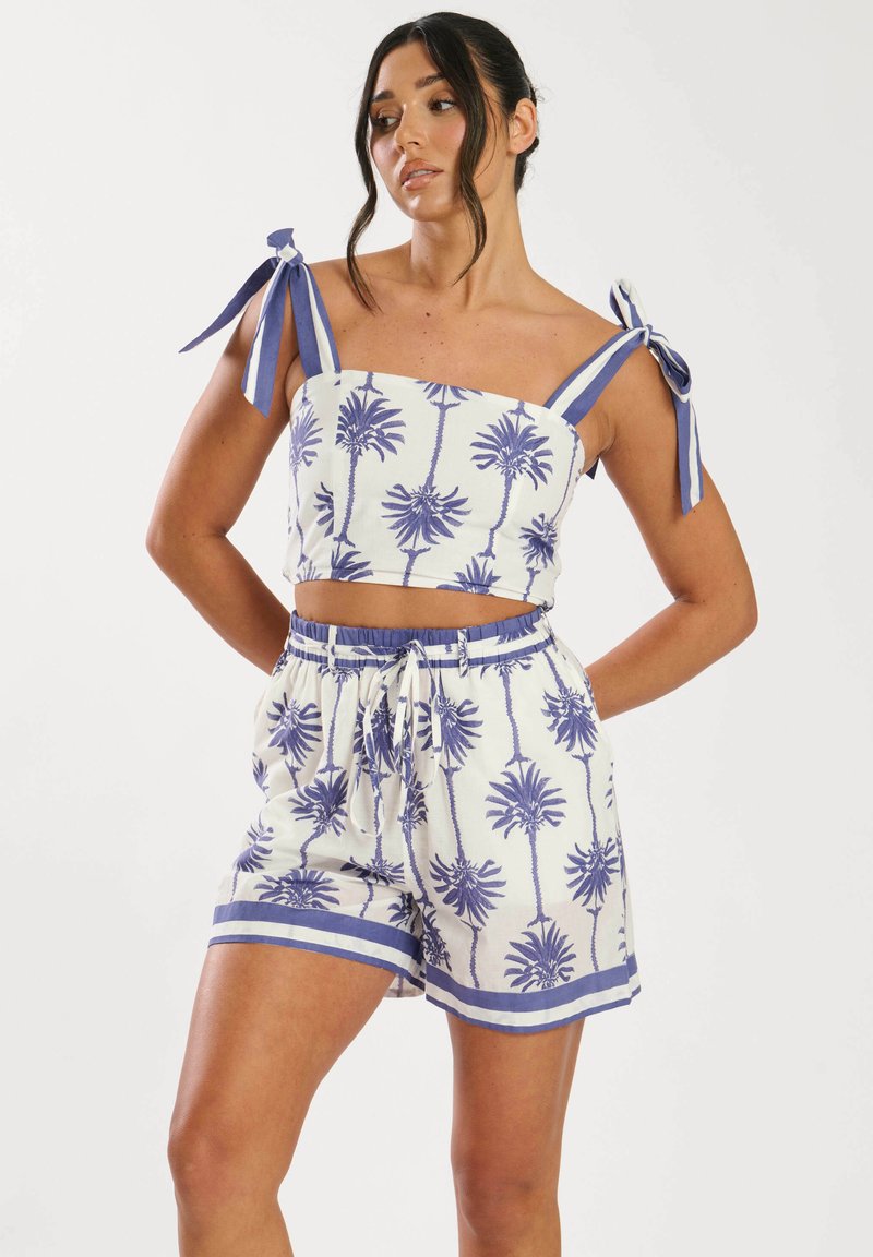 Conjunto de dos piezas que incluye un top corto y pantalones cortos elásticos. Tela blanca con estampado de palmeras azules, acentuado con tirantes anudados y cintura con cordón.