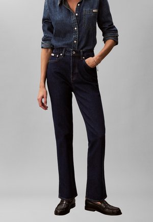 Žena oblečená v tmavomodrej denimovej košeli so zrolovanými rukávmi, zastrčenej do vysokých tmavých rifle s priamymi nohavicami a čiernych mokasín.