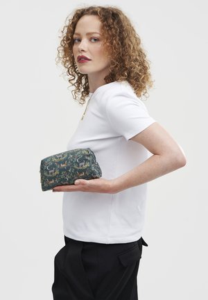Femme aux cheveux bouclés roux, vêtue d'une chemise blanche et d'un pantalon noir, tenant un sac en tissu vert foncé orné de motifs d'animaux des bois et de lunes, devant un fond uni.
