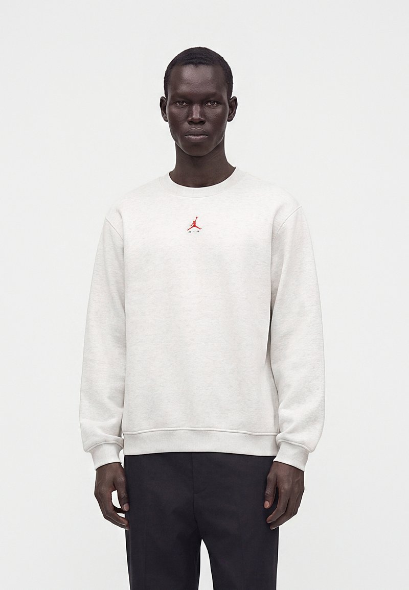 Homme portant un sweat-shirt gris clair avec logo Jordan rouge et pantalon noir, debout sur un fond blanc uni.