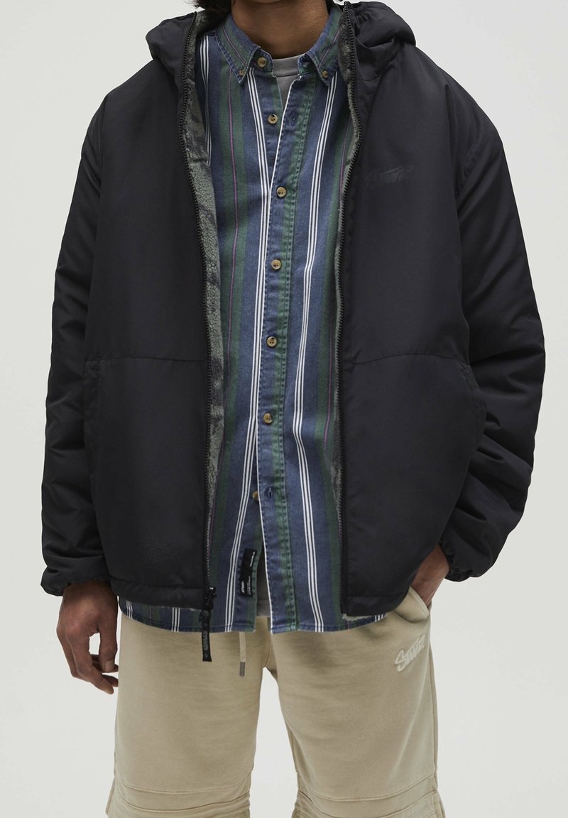 Czarna puchowa kurtka z kapturem, zapinana na zamek, noszona na niebiesko-zieloną koszulę w paski, dopełniona beżowymi szortami. Widoczne detale teksturowej tkaniny.