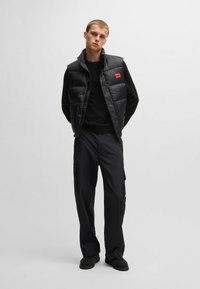 Gilet puffer nero con colletto alto, una toppa rossa con logo, indossato sopra una maglietta nera a maniche lunghe e abbinato a pantaloni cargo scuri.