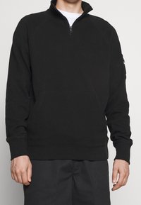Man in een zwarte kwart-rits sweatshirt met geribde mouwuiteinden en een enkele ritssluiting pocket op de linker mouw.