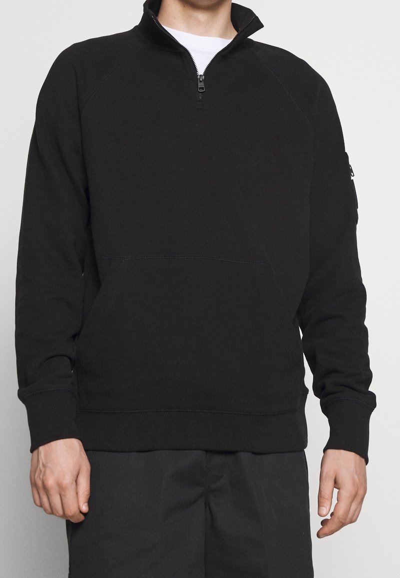 Man in een zwarte kwart-rits sweatshirt met geribde mouwuiteinden en een enkele ritssluiting pocket op de linker mouw.