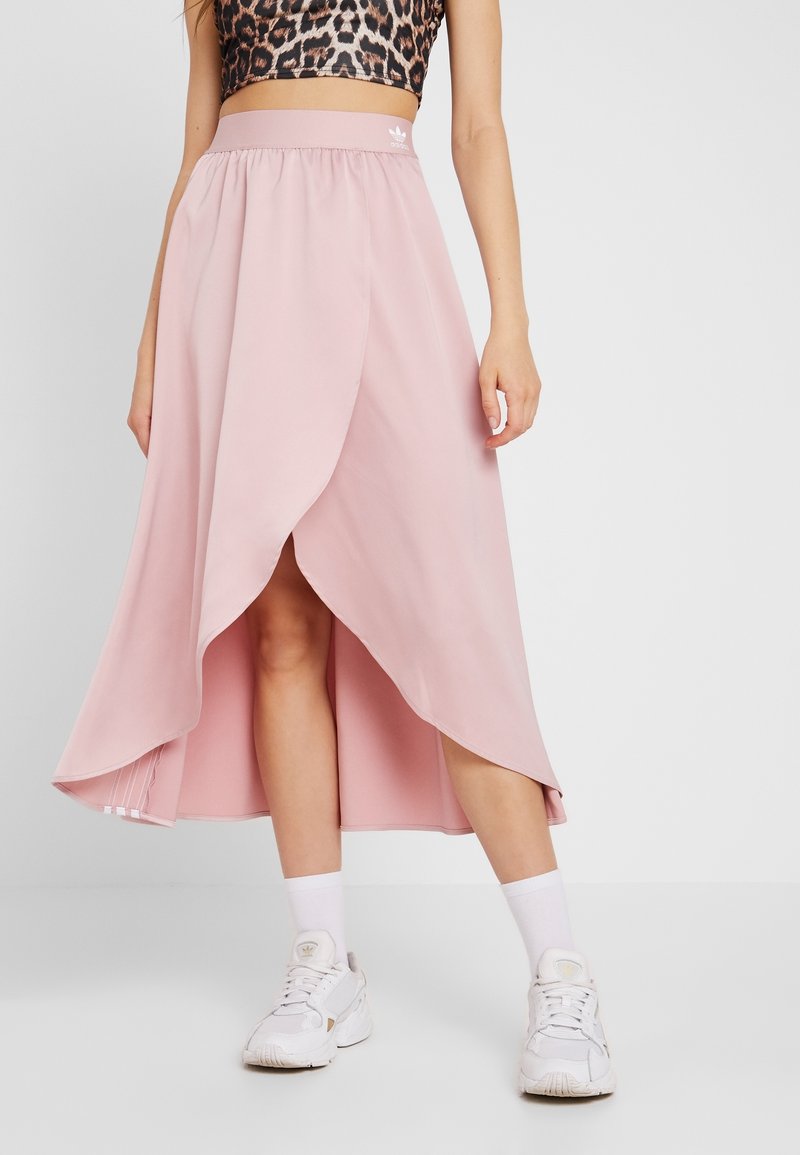 Rosa knälång kjol med asymmetrisk fåll och elastisk midja. Slätt tyg utan synliga mönster, kombinerad med vita sneakers.