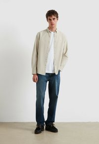 Lichtbeige overhemd met knopen, wit T-shirt eronder, blauwe spijkerbroek en zwarte schoenen met gestructureerde zolen, staand tegen een neutrale achtergrond.