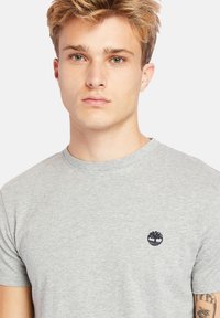 Grijze katoenen t-shirt met een ronde hals, korte mouwen en een klein donker logo geborduurd op de borst. Zachte textuur en casual pasvorm.