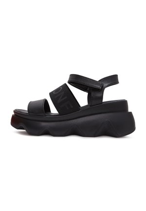 Sandale plateforme noire avec semelle épaisse en zigzag, deux larges brides dont une avec texte logo, et une bride à la cheville avec fermeture Velcro.