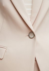 Blazer beige avec un seul bouton rond fermé sur un haut rayé crème et blanc.