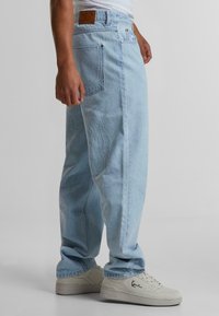 Jeans din denim de culoare albastră deschisă cu cusături albe în formă de flacără, având o etichetă din piele la talie și două buzunare la spate. Asortați cu adidași albi.