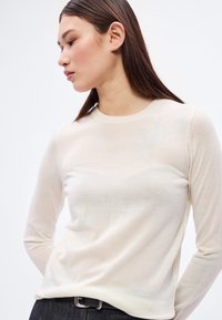 Pulover crem cu mâneci lungi, realizat dintr-un material tricotat moale, cu decolteu rotund și siluetă ajustată. Design simplu, fără modele.